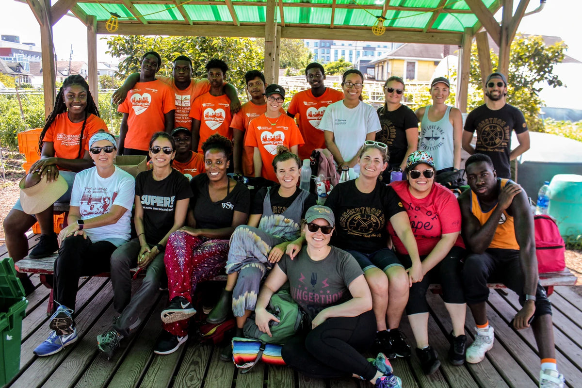 A group of twenty people posing together on a wooden deck under a green-roofed shelter. Many are wearing matching bright orange t-shirts with a white heart graphic and the word 'Cruel'.