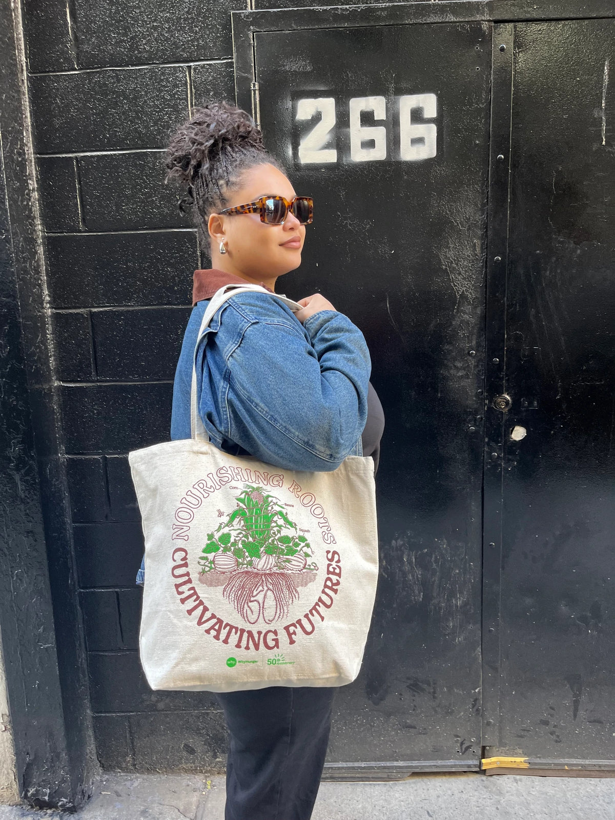 A woman wearing tortoiseshell sunglasses and a denim jacket stands in front of a black brick wall, carrying a cream canvas tote bag with a green and red graphic that reads 'Cultivating Futures' and 'November 2025 Harvest'.