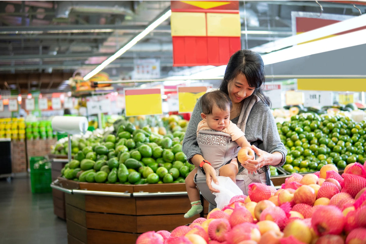 A woman in a gray cardigan holds a baby while shopping in a supermarket produce section, surrounded by fresh green limes and red apples.