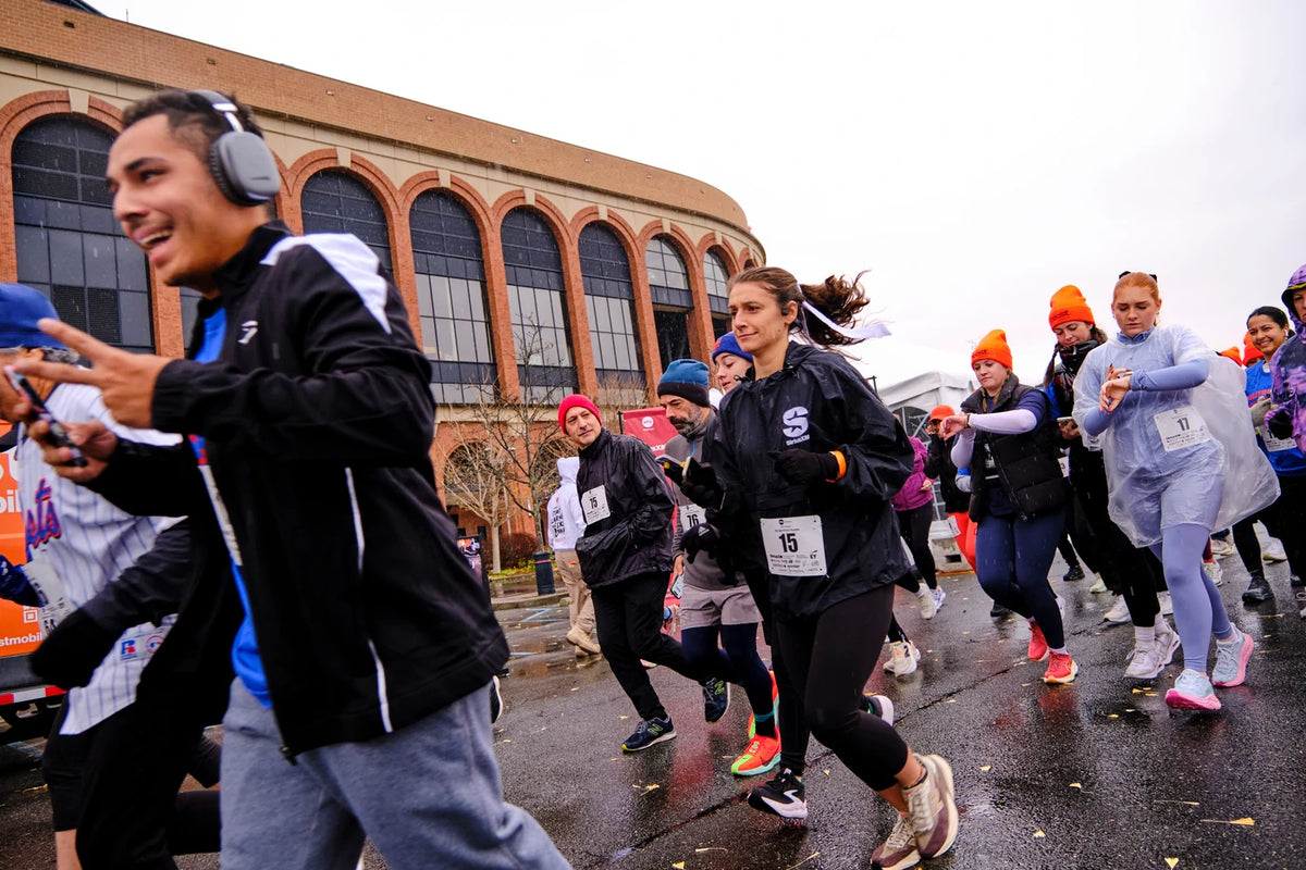 Runners on a wet asphalt road during a race, with a red-brick stadium and arched windows in the background; a smiling man with headphones and a peace sign in the foreground, and a woman in a black jacket with bib number 15 beside him.